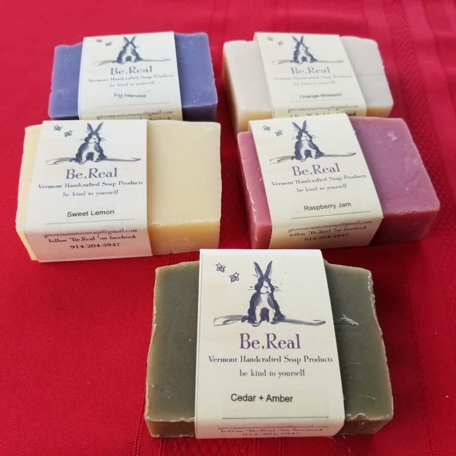 A collection of various handcrafted soap bars from Be.Real, labeled with scents like Sweet Lemon, Raspberry Jam, and Cedar + Amber.