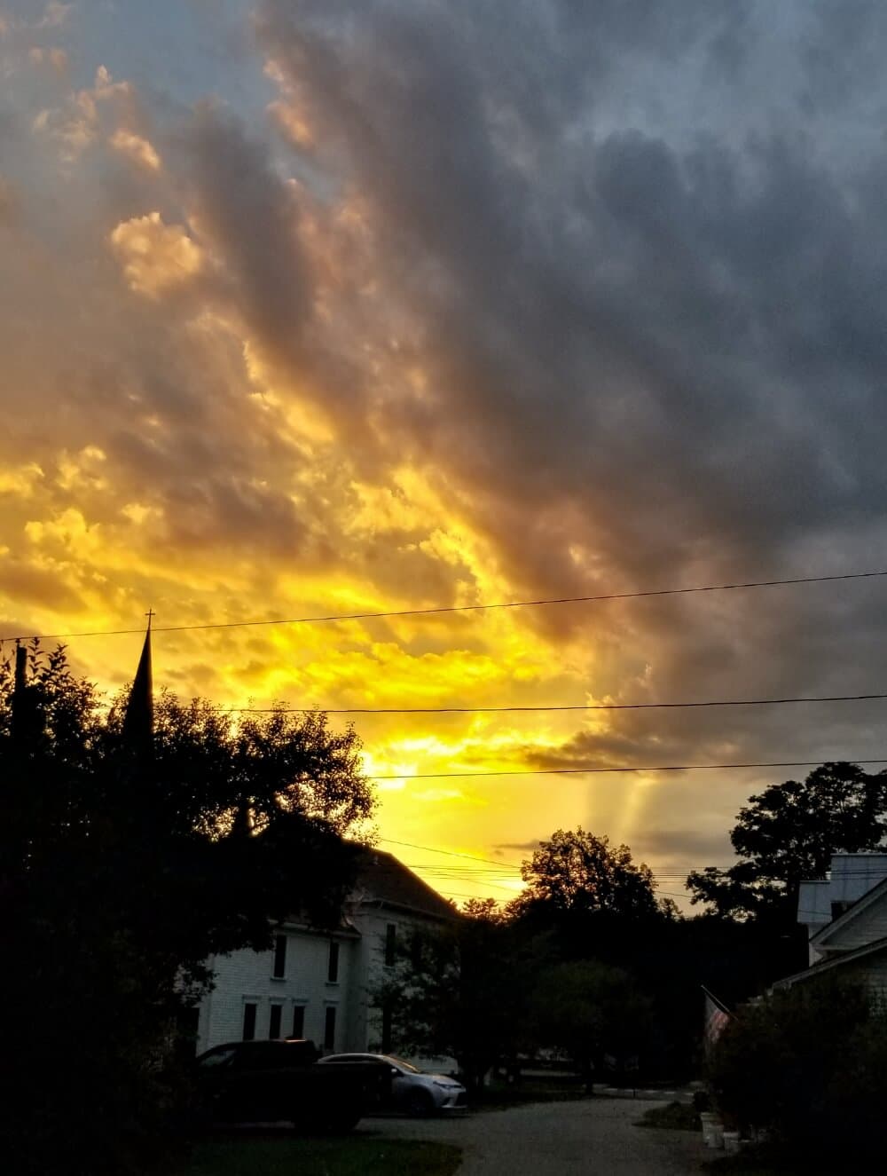 Sunset with golden hues and dramatic clouds behind a silhouette of buildings and trees.