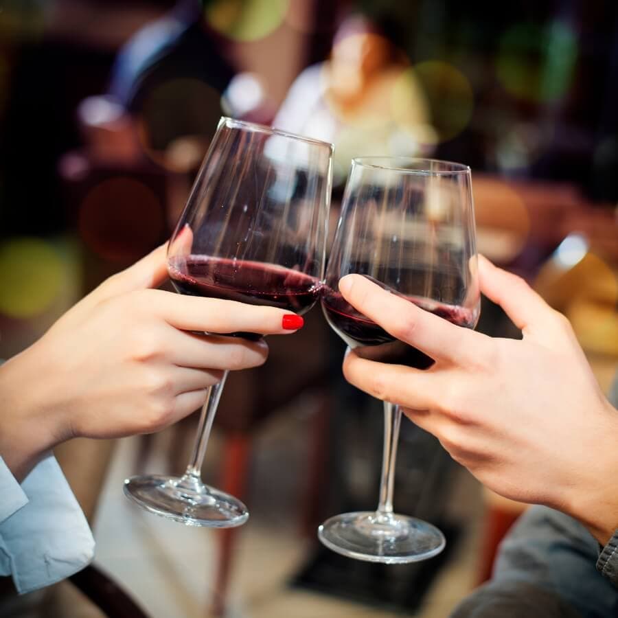 Two hands clink wine glasses filled with red wine in a cozy setting.