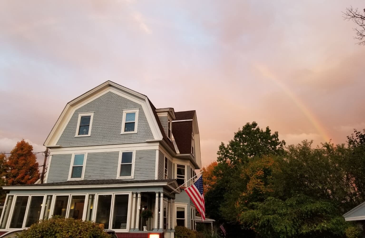 A gray house with American flag in front and a rainbow against a sunset sky.