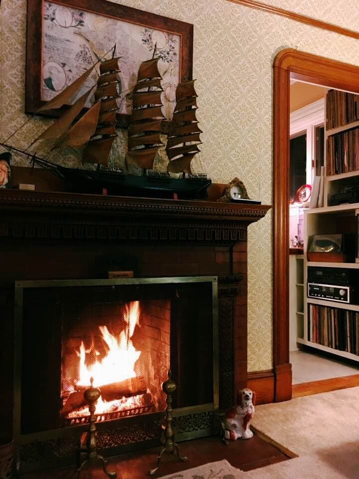 A cozy living room featuring a fireplace where a fire is crackling, surrounded by vintage decor, a ship model, and a record player.