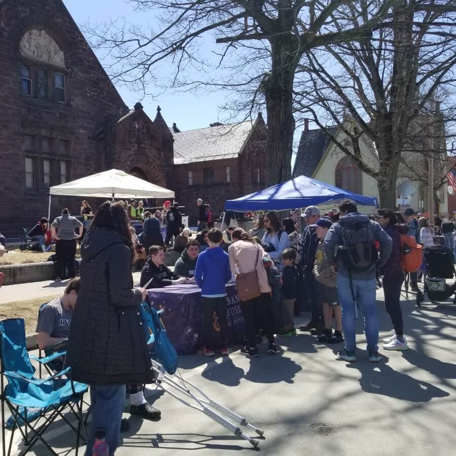 A crowded outdoor event with tents and people gathered around tables in front of historic buildings on a sunny day.