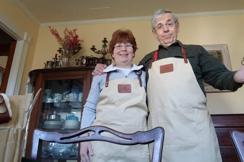 A smiling couple in matching aprons pose together in a cozy dining room.