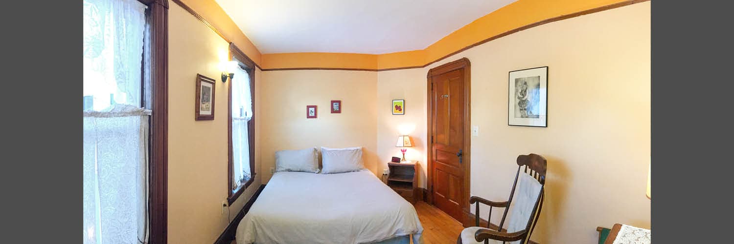 A cozy bedroom featuring a bed, rocking chair, and framed artwork on light-colored walls.