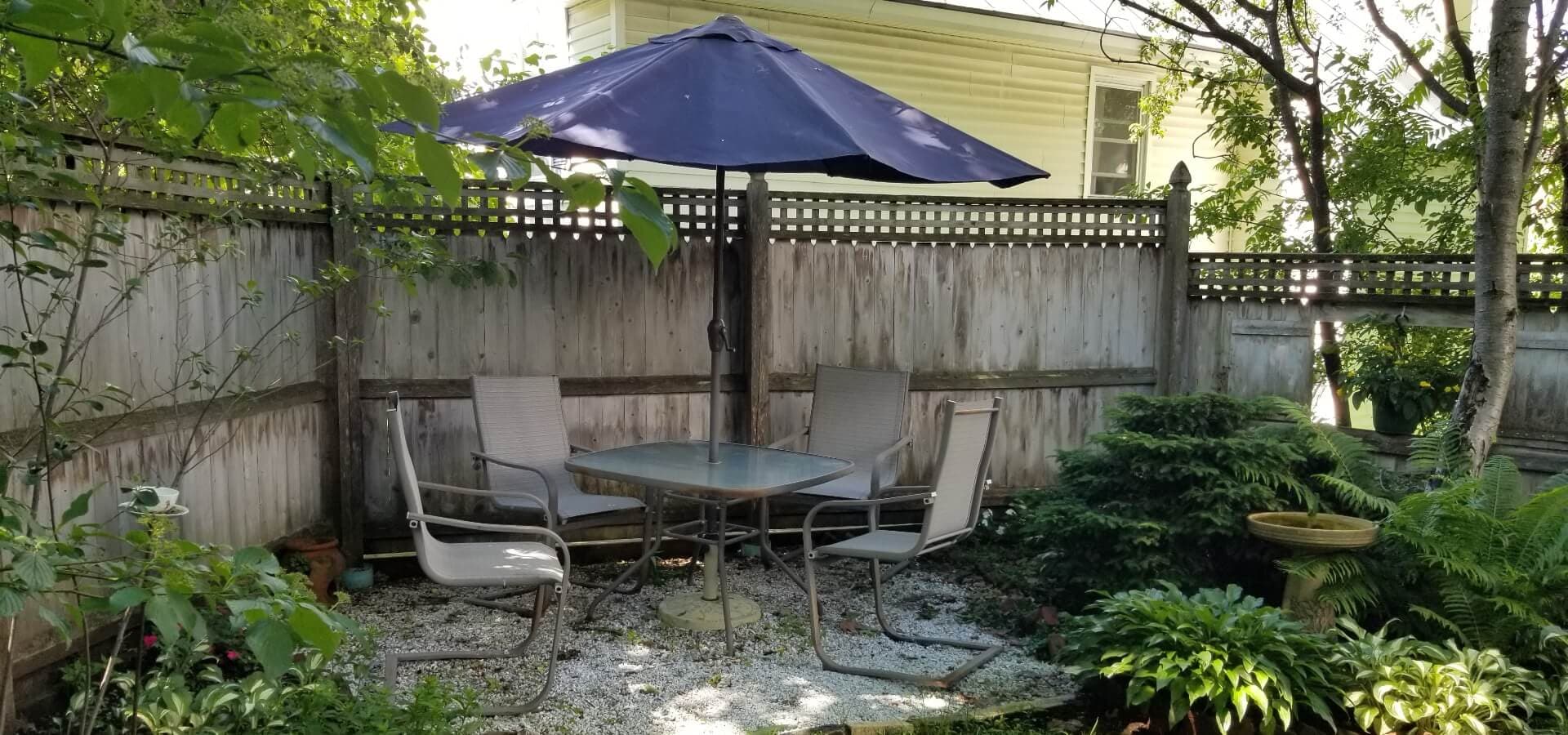 A shaded outdoor seating area with a table and chairs surrounded by greenery and a wooden fence.
