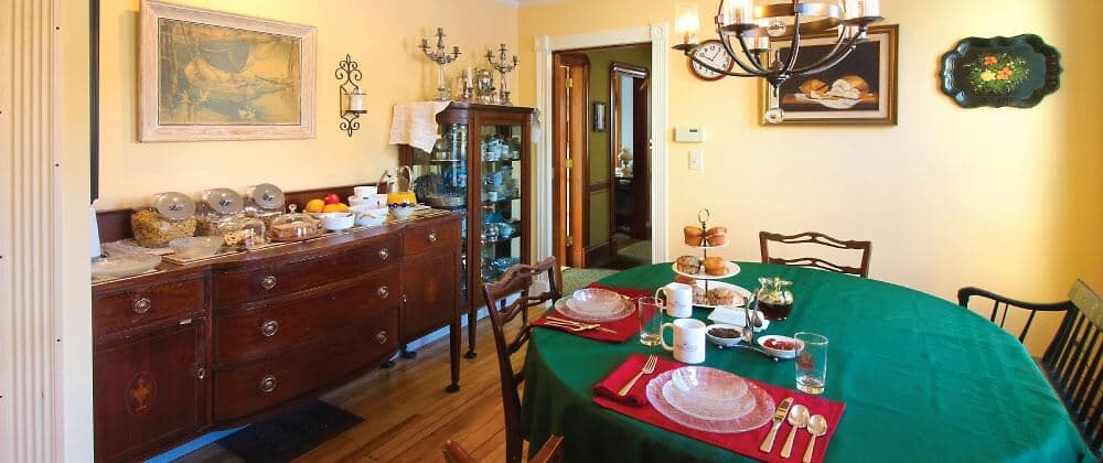 A cozy dining room set for a meal, featuring a green tablecloth, plates, silverware, and a sideboard with assorted food items.