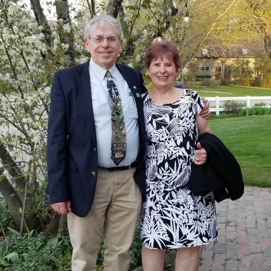 A smiling couple stands together outdoors in front of flowering trees, dressed for a special occasion.